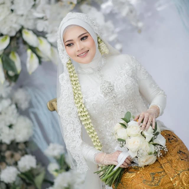 Pengantin berhijab putih tersenyum sambil memegang buket putih dan hijau