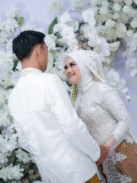 Pasangan pengantin berbusana putih saling memandang di depan dekorasi bunga